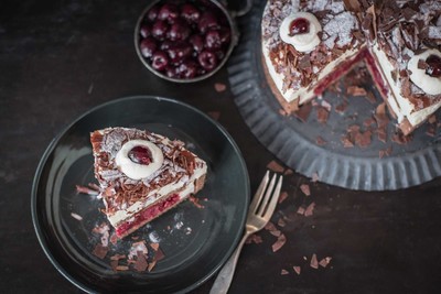 Auf einem dunklen Teller liegt ein großes Stück Schwarzwälder Kirschtorte. Rechts neben dem Teller steht der Rest der Torte. Die Torte ist mit Sahne, Schokoraspeln und Kirschen dekoriert.