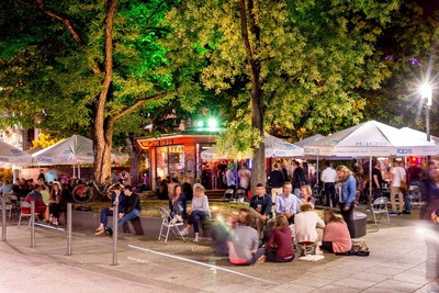 Auf einem Platz treffen sich viele junge Menschen. Sie sitzen teilweise auf Mauern und auf dem Boden. Im Hintergrund sind Stände aufgebaut an denen es etwas zu Essen und Trinken gibt.